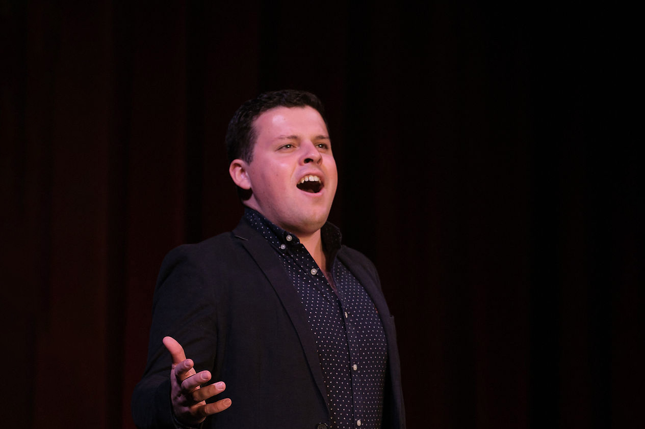 A male student, wearing a dark patterned shirt and a blazer, singing on stage with red curtains behind him.